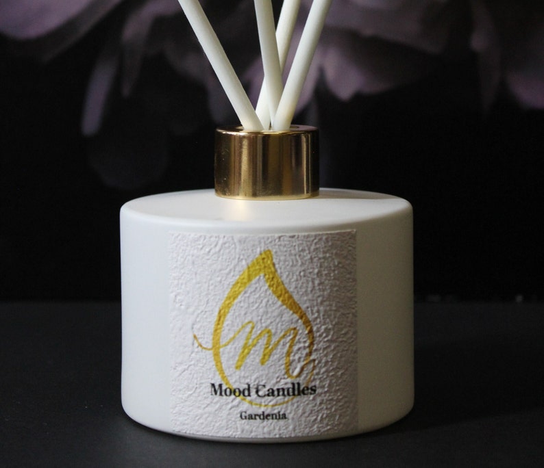 May include: White diffuser bottle with a gold lid and sticks. The bottle has a label with a gold drop and the text "Mood Candles Gardenia".