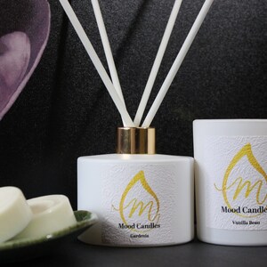 May include: White candle jars with gold lettering and a white diffuser bottle with gold accents. The candle jars have the text "Mood Candles" and the scents "Vanilla Bean" and "Gardenia". The diffuser bottle has the text "Mood Candles" and the scent "Gardenia".