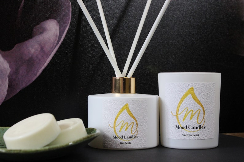 May include: White candle jars with gold lettering and a white diffuser bottle with gold accents. The candle jars have the text "Mood Candles" and the scents "Vanilla Bean" and "Gardenia". The diffuser bottle has the text "Mood Candles" and the scent "Gardenia".