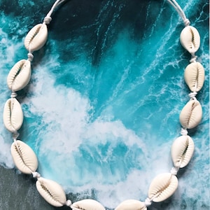 Può includere: Collana di conchiglie di cauri color crema infilate su un cordoncino bianco. Le conchiglie sono distanziate uniformemente e annodate in posizione. La collana è esposta su uno sfondo blu e bianco a tema oceanico.