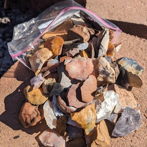 May include: A plastic bag filled with a variety of colorful, chipped, and broken rocks. The rocks are mostly brown, tan, and white, with some hints of pink and gray. The rocks are sitting on a brown, textured surface.