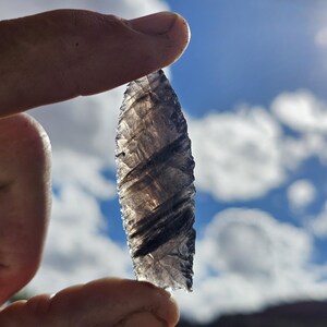 May include: A translucent, gray arrowhead with dark, diagonal striations is held between fingers against a backdrop of a blue sky with white clouds. The arrowhead is a teardrop shape, with a sharp point.