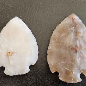 May include: Two stone arrowheads. One is a light cream color, the other is a light brown with red markings. Both have a teardrop shape with a notched base. The arrowheads are likely hand-crafted and are a light color.