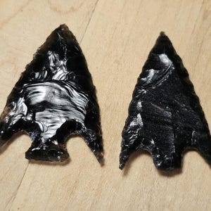 May include: Two obsidian arrowheads with a triangular shape and notched base. The arrowheads are a deep black color with a glossy, reflective surface. The edges are sharp and well-defined, showcasing the craftsmanship.
