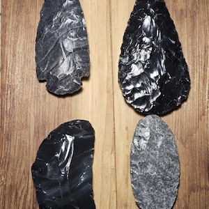 May include: Four ancient obsidian and stone artifacts are displayed on a wooden surface. Two are dark, glossy arrowheads, one with a notched base. The other two are a black, irregular shard and a gray, oval-shaped stone.