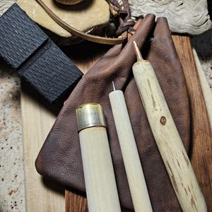 May include: A collection of handcrafted leatherworking tools and accessories. The image features a brown leather pouch, wooden handles with metal tips, a black textured block, and a stone with a leather strap. The items are arranged on a wooden cutting board.