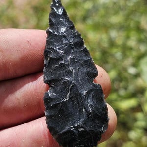 May include: A black obsidian arrowhead with a sharp, pointed tip and a textured surface. The arrowhead is held in a hand against a blurred green background, showcasing its craftsmanship and historical significance.