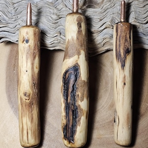 May include: Three handcrafted wooden bottle stoppers with copper-colored tops. The wood has natural grain patterns and knots, with variations in color from light tan to dark brown. Each stopper is unique in shape and wood grain.