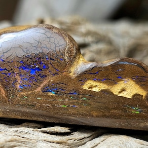 May include: A polished, natural opal gemstone with a brown base color and vibrant blue, green, and gold flashes. The opal has a unique, organic shape and a glossy surface, showcasing its internal patterns and colors.