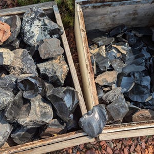 May include: Two wooden crates filled with rough, dark obsidian stones. One crate contains a large, reddish-brown rock. The obsidian pieces vary in size and shape, with a shiny, reflective surface. The crates sit on a bed of small, reddish-brown pebbles.