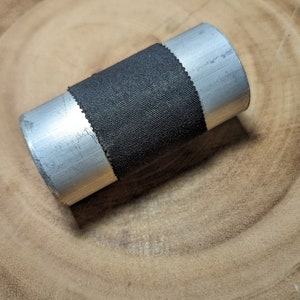 May include: A cylindrical metal object with a black fabric band wrapped around its center. The metal has a brushed appearance. The object is resting on a wooden surface with a circular grain pattern.