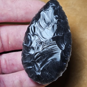 May include: A dark obsidian arrowhead with a teardrop shape. The surface of the stone has a glossy, reflective quality, with visible patterns and textures. The arrowhead is held in a hand, showcasing its size and form.