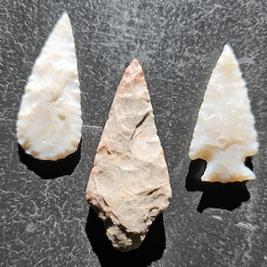 May include: Three ancient arrowheads. Two are white, and one is tan with a reddish tip. The arrowheads are made of stone and have sharp edges. They are arranged on a dark, textured surface.