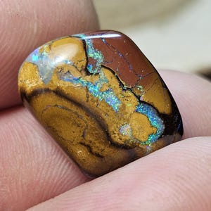 May include: A polished boulder opal gemstone featuring a smooth, rounded rectangular shape. The stone displays a rich, earthy brown ironstone matrix contrasted by vibrant, iridescent flashes of blue and green light across its surface.