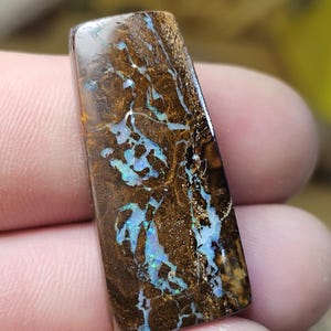 May include: A rectangular boulder opal gemstone featuring a dark brown ironstone matrix with vibrant, iridescent blue and green flashes of color running through the natural fissures and veins of the polished stone surface.