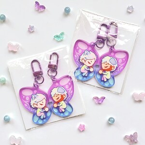 May include: Two purple and pink butterfly-shaped keychains with cartoon characters on them. The keychains are attached to silver key rings.