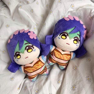 以下が含まれることがあります： 紫色の髪、ピンクの花のアクセント、詳細な顔の特徴を持つ2つのぬいぐるみ人形。オレンジとピンクの色合いの模様入りのトップスを着ており、腕と脚には青とピンクのアクセントがあります。人形は白い背景に置かれています。