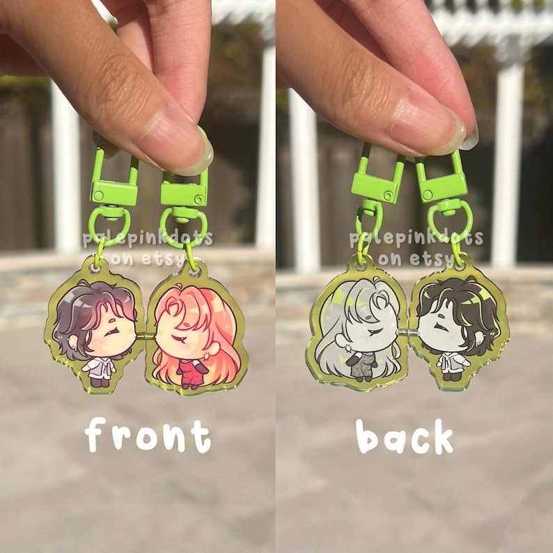 May include: Double-sided acrylic keyrings with anime-style characters. The front shows characters with pink and brown hair, the back with white and brown hair. The keyrings have green clasps. Text on the keyrings reads 'front' and 'back'.