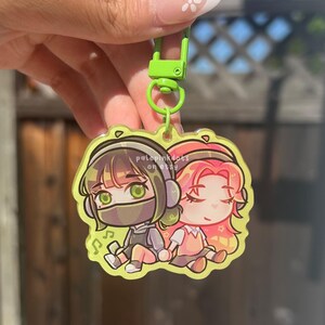 May include: A clear acrylic keychain with two anime-style characters wearing headphones, one in grey and the other in pink. The keychain has a lime green clasp and ring. The text "pale pink dots on etsy" is visible.