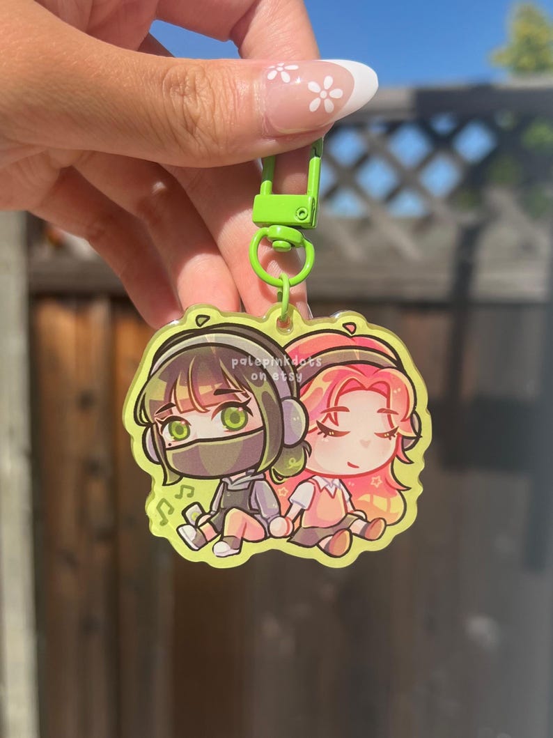 May include: A clear acrylic keychain with two anime-style characters wearing headphones, one in grey and the other in pink. The keychain has a lime green clasp and ring. The text "pale pink dots on etsy" is visible.