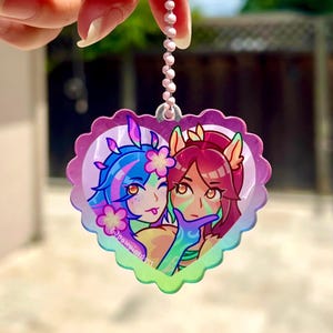 May include: A heart-shaped charm with a pink, purple, and blue iridescent finish. The charm features a cartoon image of two female characters with blue and red hair, holding hands and smiling. The charm is attached to a silver chain.