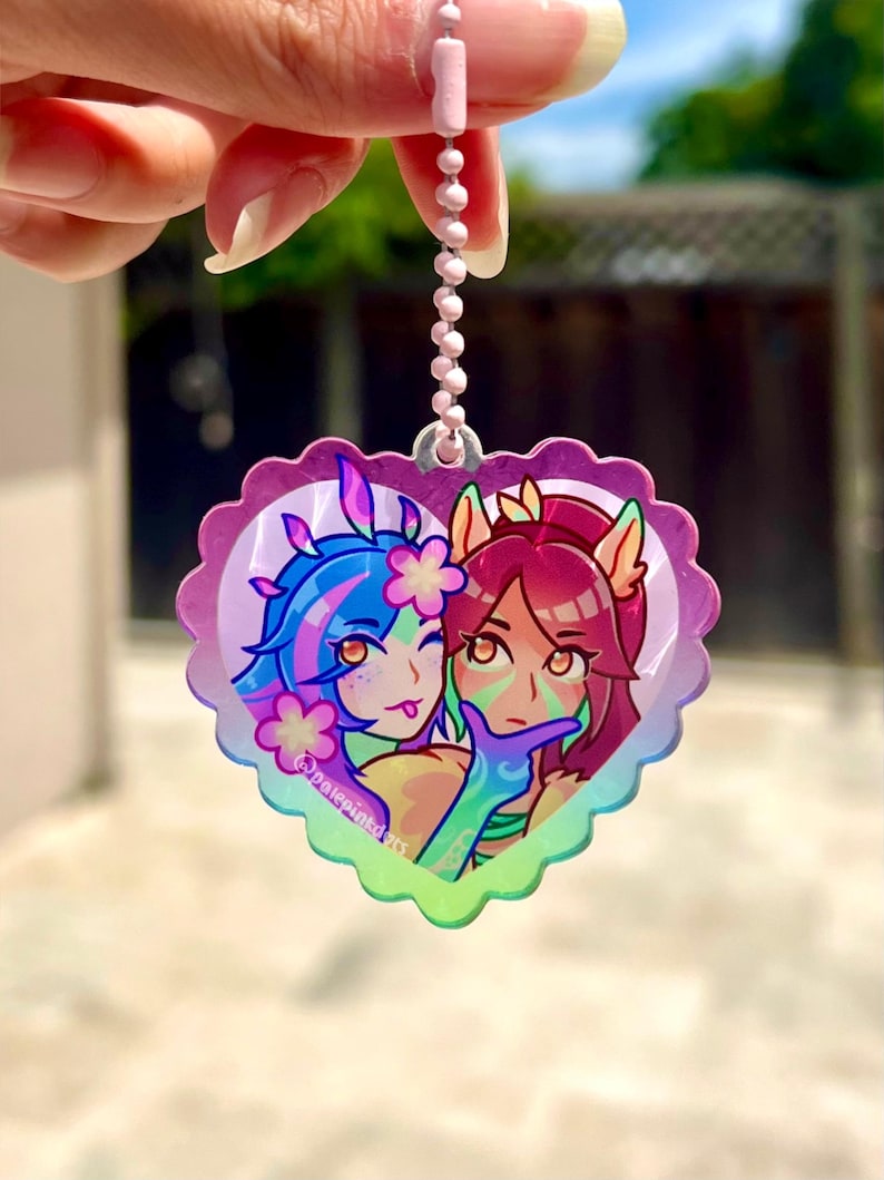 May include: A heart-shaped charm with a pink, purple, and blue iridescent finish. The charm features a cartoon image of two female characters with blue and red hair, holding hands and smiling. The charm is attached to a silver chain.