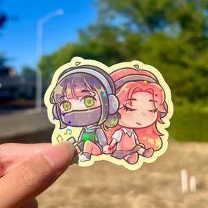May include: A vibrant sticker depicting two anime-style figures holding hands. One character has black hair and a face covering, the other has pink hair. Both are wearing headphones and seated. The sticker has a yellow border.