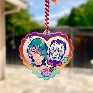 May include: A heart-shaped keychain with a colorful design featuring two anime characters. The characters are a woman with blue hair and a man with a purple and white mask. The keychain is made of clear acrylic and has a rainbow gradient effect.