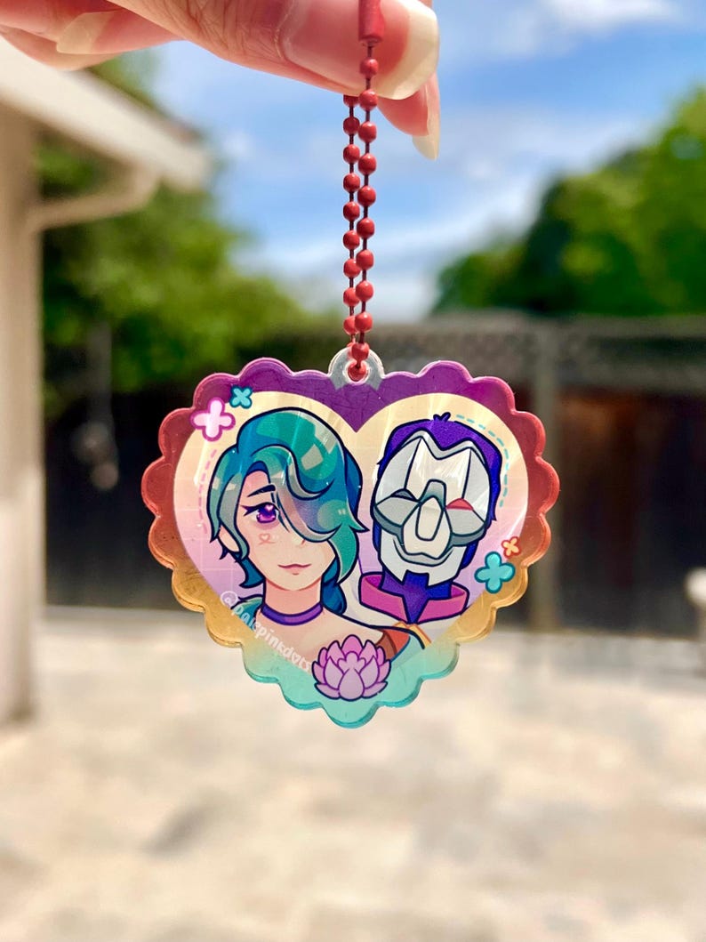 May include: A heart-shaped keychain with a colorful design featuring two anime characters. The characters are a woman with blue hair and a man with a purple and white mask. The keychain is made of clear acrylic and has a rainbow gradient effect.