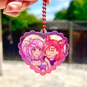 May include: A pink heart-shaped keychain with a cartoon image of two anime characters. The characters are a girl with pink hair and a girl with red hair. The keychain has a red chain.