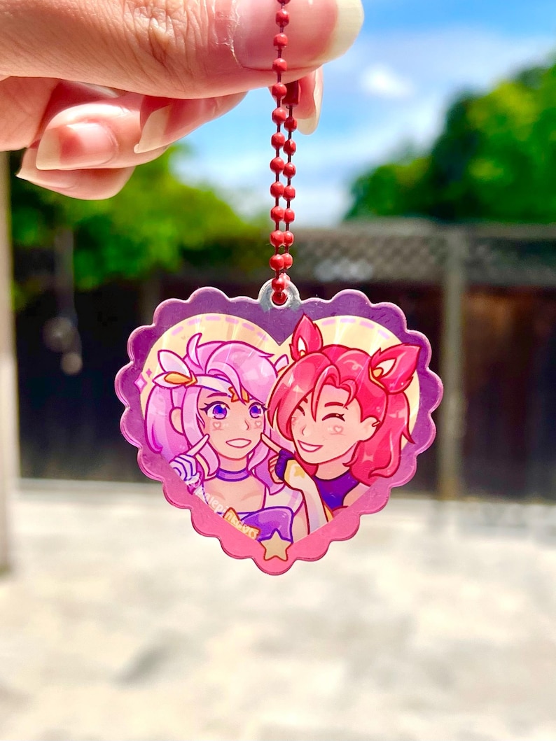 May include: A pink heart-shaped keychain with a cartoon image of two anime characters. The characters are a girl with pink hair and a girl with red hair. The keychain has a red chain.