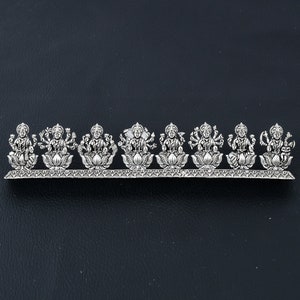 May include: A silver-toned metal decorative piece featuring eight Hindu deities, each seated on a lotus flower. The deities are arranged in a row on a decorative base.
