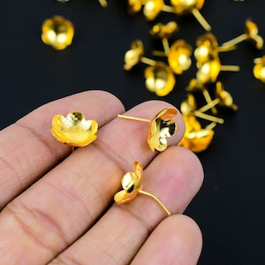 Puede incluir: Primer plano de una mano sosteniendo adornos de metal de color dorado en forma de flor con un pequeño alfiler en la parte posterior. Los adornos son perfectos para agregar un toque de elegancia a las manualidades y las joyas.