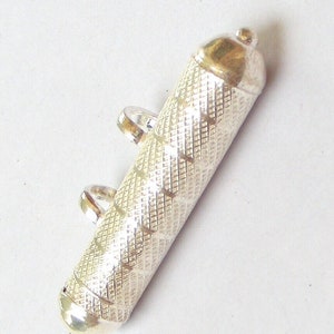 May include: A silver-colored cylindrical pendant with a textured, diamond-patterned surface. The pendant has a rounded top and bottom, with two small loops for attaching a chain. The top and bottom have a gold-colored finish.