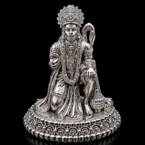 Puede incluir: Una figura metálica de tono plateado de una deidad arrodillada sobre una base circular ornamentada. La figura lleva una corona, collares superpuestos y brazaletes, con una mano levantada en gesto de bendición y la otra cerca de una maza.