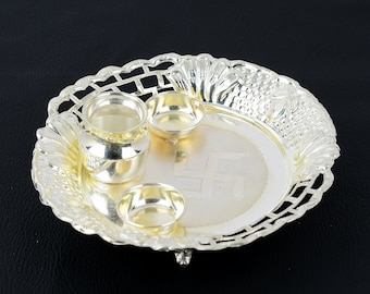 May include: A silver-coloured metal plate with intricate designs, featuring three small bowls and a swastika symbol in the centre. The plate has three feet and is perfect for religious ceremonies.