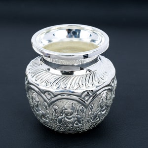 May include: A silver-colored, ornate pot with a wide rim and detailed carvings. The pot features a bulbous body with intricate relief designs of figures and floral patterns. The polished surface reflects light, highlighting the detailed craftsmanship. The pot sits on a dark surface.