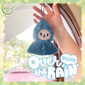 May include: A blue crochet doll with a hooded cape. The doll has a white face with black eyes and a small mouth. The image includes the text "Over the Rain" and "SKZOO".