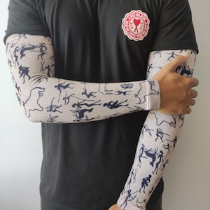 Pode incluir: Uma pessoa vestindo uma camiseta preta com um logotipo de coração vermelho que diz "AMI PARIS" e um par de aquecedores de braço bege com um padrão de figuras de pau preto e branco.
