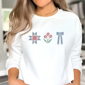 May include: White sweatshirt with embroidered designs. The designs include a starburst, three pink flowers, and a blue bow. The sweatshirt is a long-sleeved crew neck style.