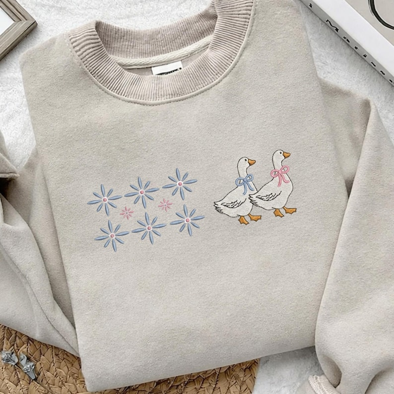 May include: A beige sweatshirt with embroidered details. The design features two geese with bows and a cluster of blue and pink flowers. The sweatshirt has a ribbed neckline and cuffs, and a relaxed fit.