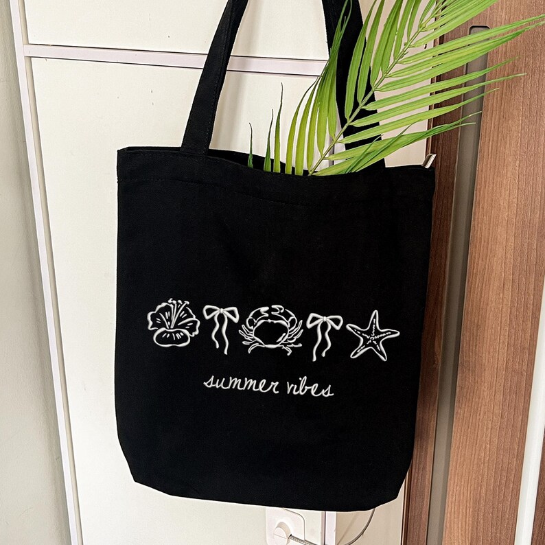 K&ouml;nnte beinhalten: Schwarze Tragetasche mit wei&szlig;en, gestickten Sommermotiven, darunter eine Blume, Schleifen, eine Krabbe und ein Seestern. Die Worte "summer vibes" sind darunter in Schreibschrift geschrieben. Gr&uuml;ne Palmbl&auml;tter sind im Inneren der Tasche zu sehen.