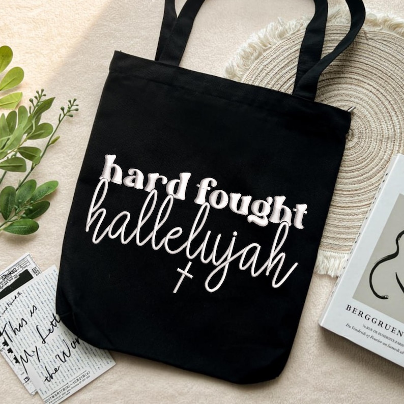 K&ouml;nnte beinhalten: Schwarze Tragetasche mit den Worten "hard fought hallelujah" in wei&szlig;er Schrift. Die Tasche hat zwei schwarze Griffe. Ein kleines Kreuz befindet sich unter dem Wort "hallelujah".