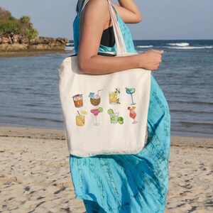 May include: A cream-colored tote bag featuring watercolor illustrations of various cocktails. The bag is held by a person on a beach, with the ocean and a rocky shoreline in the background. The cocktails include a coconut drink, margarita, and a mai tai.