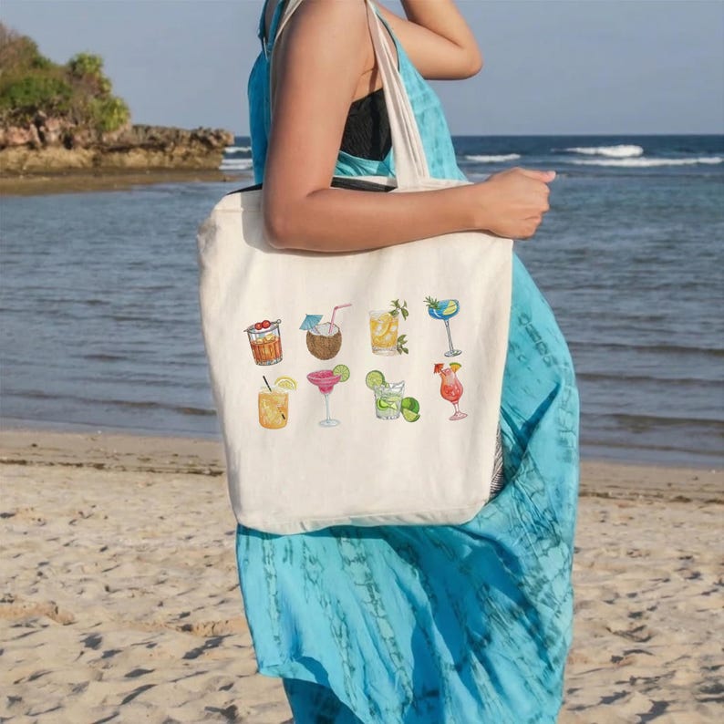 May include: A cream-colored tote bag featuring watercolor illustrations of various cocktails. The bag is held by a person on a beach, with the ocean and a rocky shoreline in the background. The cocktails include a coconut drink, margarita, and a mai tai.