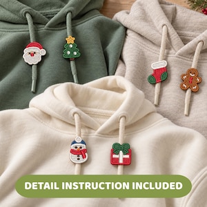 May include: Three hooded sweatshirts in olive green, beige, and cream, each featuring two thick drawstrings with embroidered holiday-themed charms. The charms include a Santa Claus, a Christmas tree, a stocking, a gingerbread man, a snowman, and a gift.