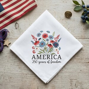 Puede incluir: Pa&ntilde;uelo blanco con un dise&ntilde;o floral en rojo, blanco y azul. El texto "AMERICA 250 years of freedom" est&aacute; impreso debajo de las flores. Una bandera americana est&aacute; en el fondo.