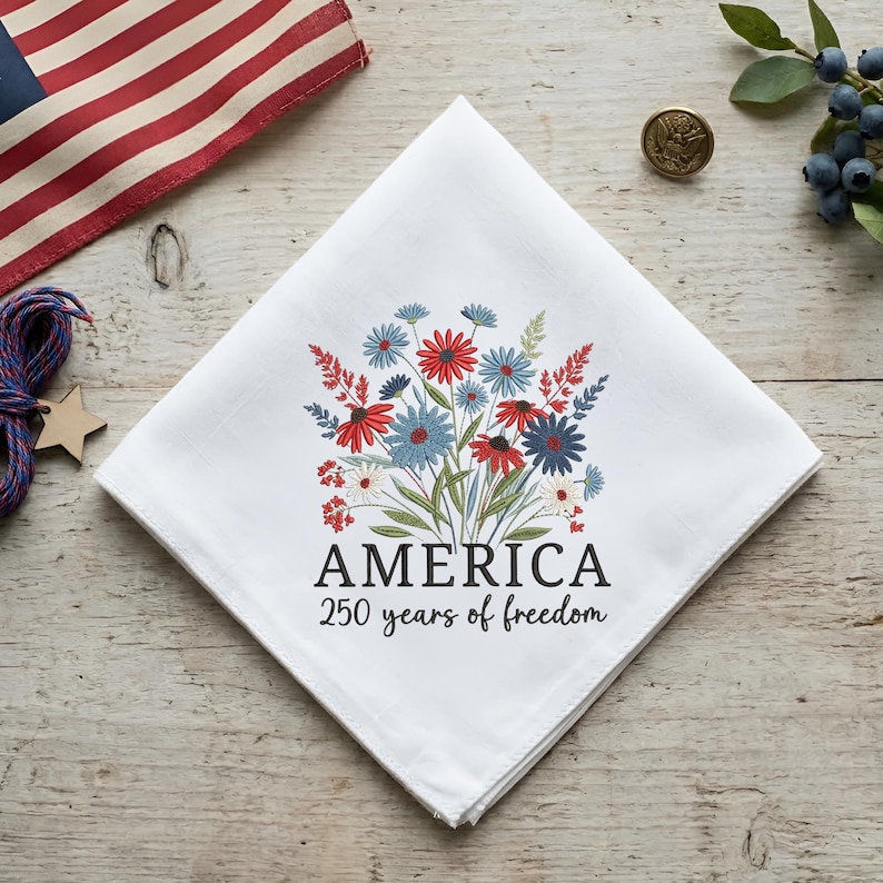 Puede incluir: Pa&ntilde;uelo blanco con un dise&ntilde;o floral en rojo, blanco y azul. El texto "AMERICA 250 years of freedom" est&aacute; impreso debajo de las flores. Una bandera americana est&aacute; en el fondo.