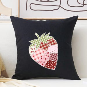 May include: Black square throw pillow featuring a patchwork strawberry design. The strawberry is composed of various fabric patterns, including gingham, polka dots, and floral prints in shades of red, pink, and green. The pillow is a decorative home accent.