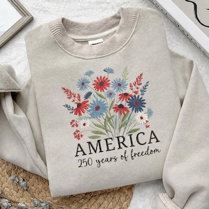 Puede incluir: Sudadera beige con un dise&ntilde;o de ramo floral en rojo, blanco y azul. La palabra "AMERICA" est&aacute; impresa debajo de las flores, con "250 years of freedom" debajo.